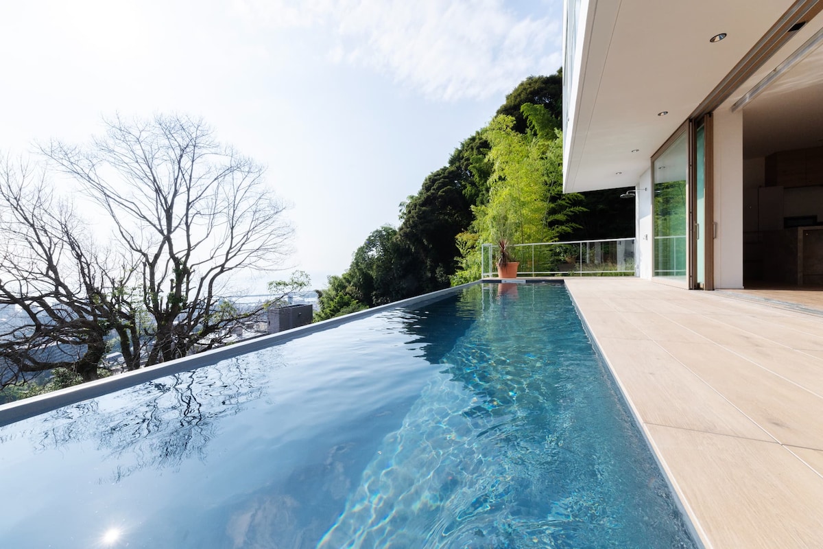 An infinity pool extends into the horizon, reflecting the clear sky and surrounding greenery. The smooth tiled edge blends seamlessly with the light wood decking, creating an inviting space. Beyond the pool, trees gently frame the landscape, enhancing the tranquil ambiance.
