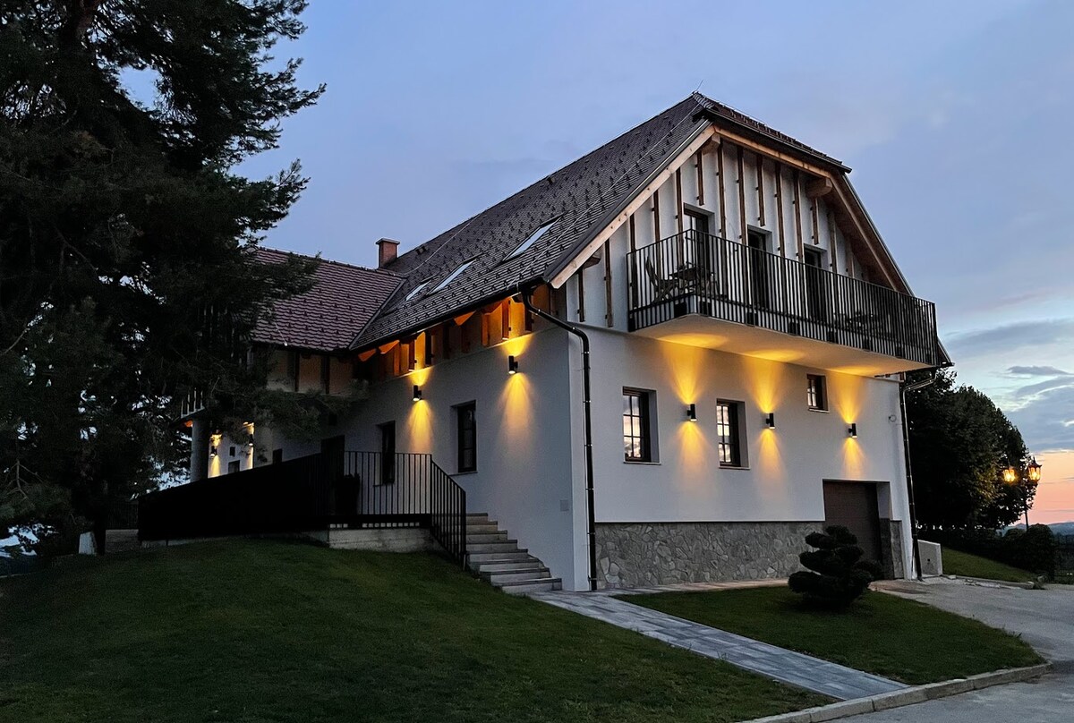 The exterior of a two-story establishment is illuminated with soft lighting, highlighting the architectural details. A spacious lawn surrounds the building, with a stone pathway leading to the entrance. A balcony extends from the upper level, enhancing the serene atmosphere of the setting.