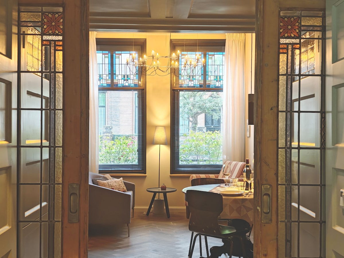 A view through ornate double doors reveals a bright living area featuring large windows adorned with stained glass. Comfortable seating is arranged near a small table, with a soft lamp providing gentle illumination. Natural light filters in, highlighting the elegant decor and inviting atmosphere.