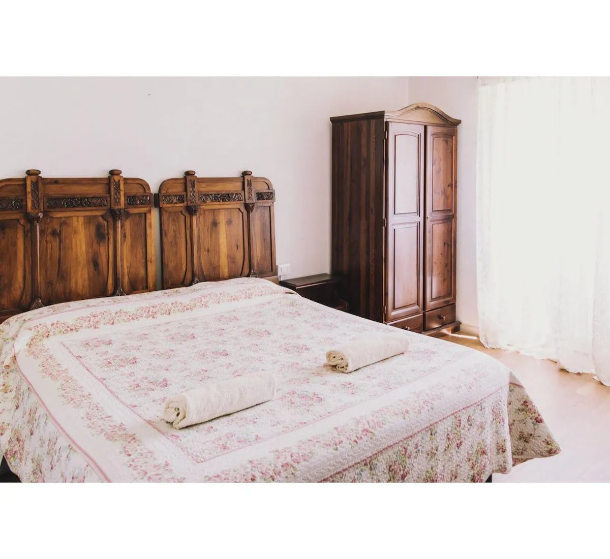 A spacious bedroom features a wooden bed frame with an intricately carved headboard, adorned with a floral patterned bedspread. Two rolled towels are placed neatly at the foot of the bed. A wooden wardrobe stands nearby, while natural light filters in through a window with sheer curtains.