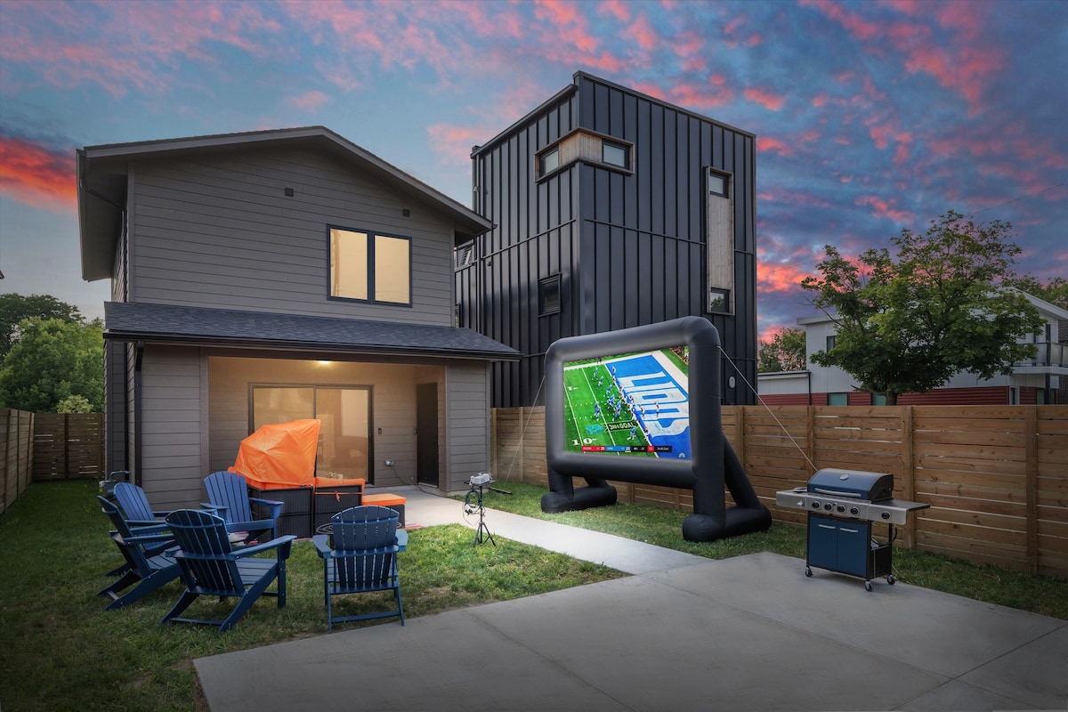 An outdoor living space features a patio with blue Adirondack chairs facing a vibrant inflatable movie screen. A BBQ grill stands nearby, with a spacious yard surrounded by a privacy fence. The dusk sky showcases a blend of pink and purple hues.