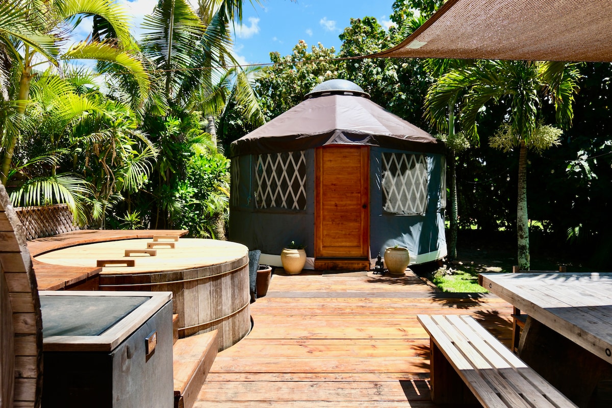 A wooden deck surrounds the yurt with lush greenery providing a natural backdrop. The entrance features a wooden door, framed by large palm leaves. An outdoor hot tub and a shared spa area are visible, inviting relaxation in a serene setting.