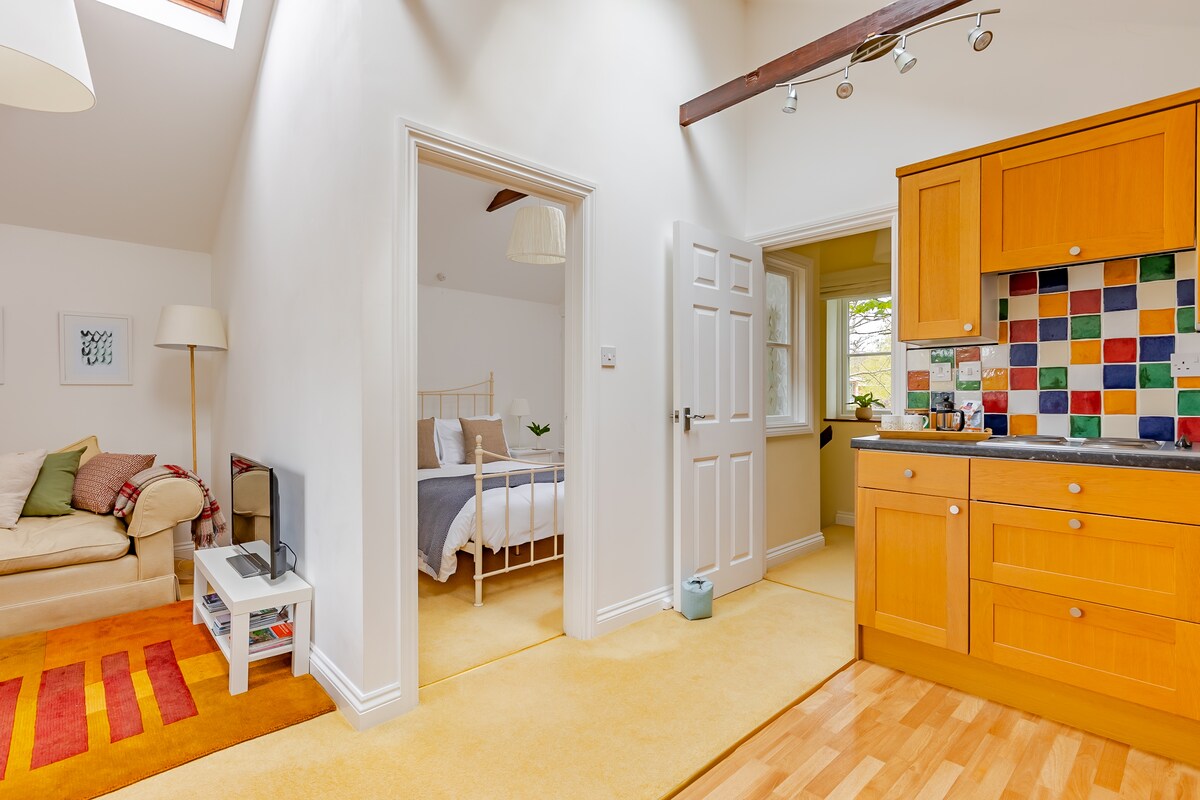 The open plan living area connects to a bright kitchen featuring colorful tiled backsplashes. A comfortable seating area with a television faces a cozy bedroom entrance. Natural light enters through skylights and windows, enhancing the inviting space. Soft yellow carpeting completes the warm atmosphere.