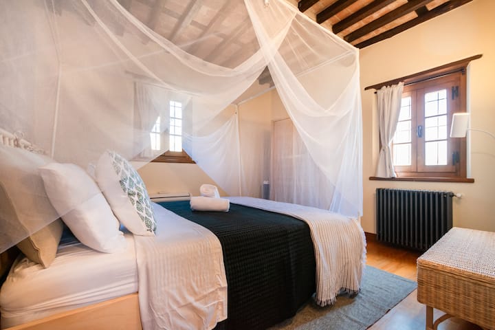 Cama tamaño queen con mosquitera y marco, baño con bañera/ducha adyacente a la habitación, armario, banco con almacenamiento, mesa auxiliar, lámpara y espejo.