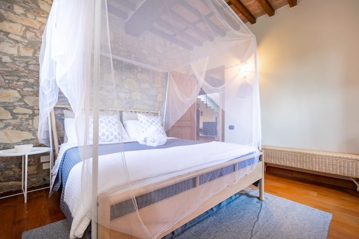 Cama tamaño queen con mosquitera y marco, baño con ducha adyacente a la habitación, armario, banco con almacenamiento, mesa auxiliar, lámpara y espejo.