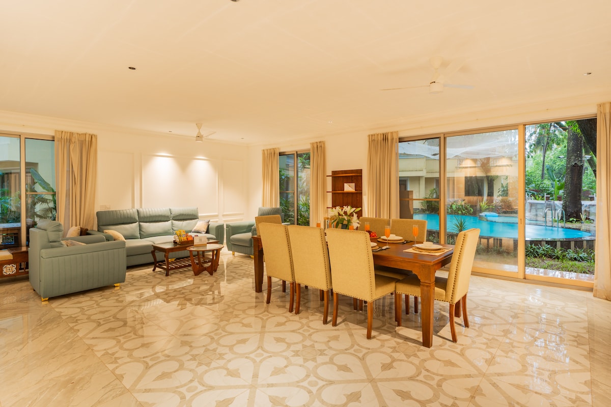 A spacious living and dining area features large glass sliding doors providing views of the pool and garden. Comfortable seating options include a three-seater sofa and a dining table for eight. Smooth, patterned floor tiles enhance the cozy, well-lit space adorned with natural light.