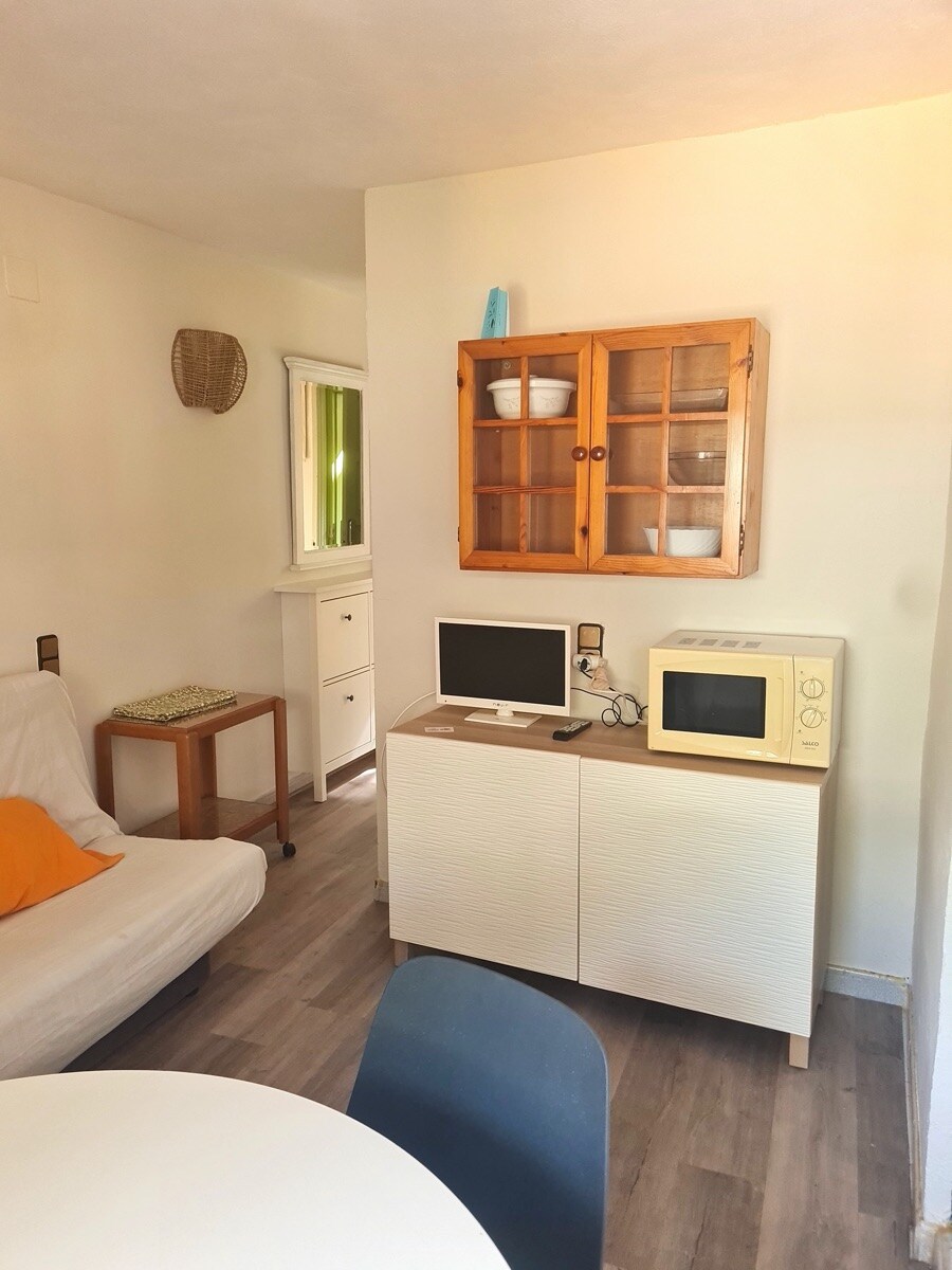 A cozy living area features a light-colored sofa bed and a round dining table with chairs. A compact white cabinet houses a microwave and a small computer monitor. Natural light filters in through a nearby window, adding brightness to the space.