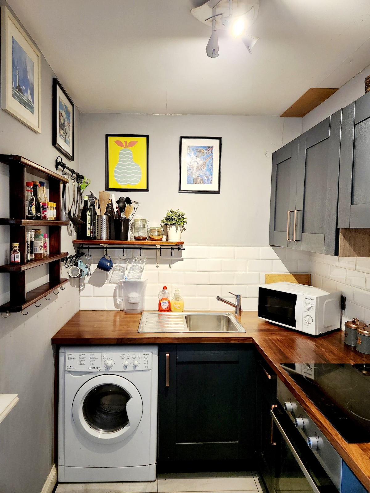 A compact kitchen is presented, featuring dark cabinetry and a wooden countertop. A washing machine is positioned next to a stainless-steel sink. Decorative shelves hold various kitchen items, and a microwave is visible. Artwork adorns the wall, adding a touch of character to the space.