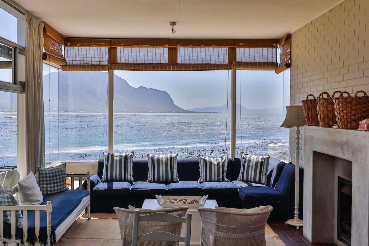 A light-filled living area features large glass windows that frame stunning views of the ocean and mountains. Comfortable seating with striped cushions faces the water, while a cozy fireplace adds warmth. Natural light enhances the serene ambiance throughout the space.