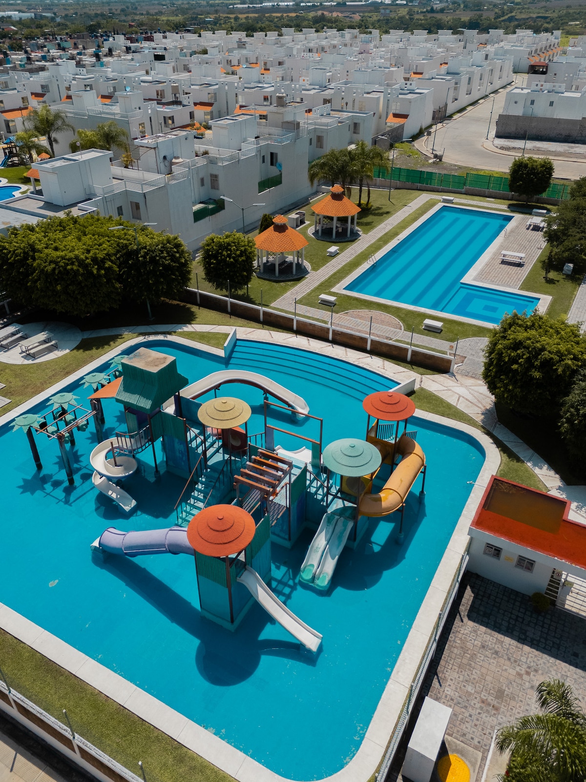 An aerial view captures the vibrant water park, featuring colorful slides and play structures set in a large, bright blue pool. Adjacent is a semi-Olympic pool, bordered by well-kept greenery and lounge areas. The surrounding residential complex is visible in the background.