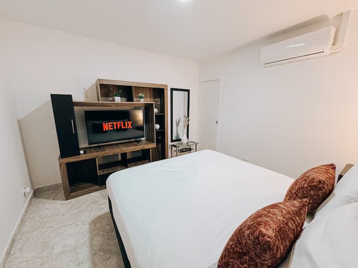 Habitación moderna con cama queen, cojines decorativos en tonos terracota y aire acondicionado. Frente a la cama hay un centro de entretenimiento en madera con TV y acceso a Netflix. Incluye espejo de cuerpo entero y mesa lateral con florero.