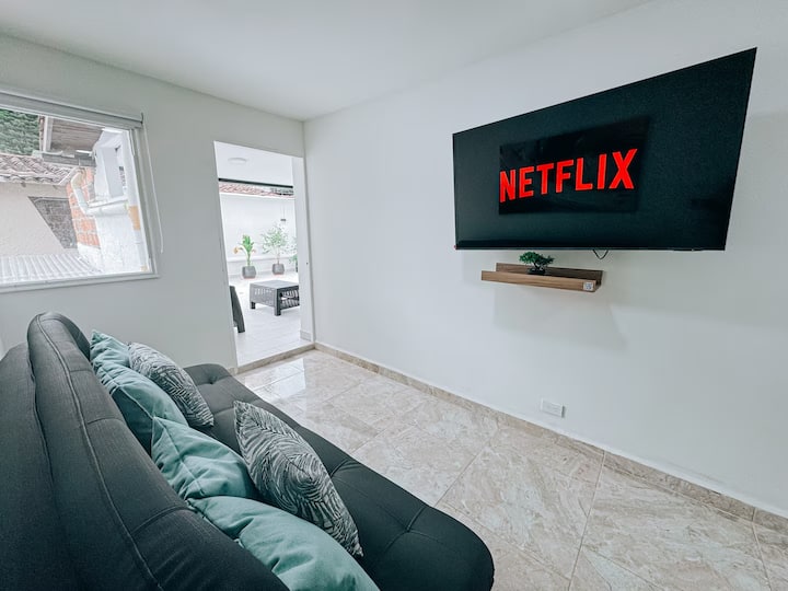 Sala con estilo minimalista y moderno, con sofá negro acolchado decorado con cojines en tonos grises y verdes. Frente al sofá, un televisor de pantalla plana con acceso a Netflix montado en la pared y repisa flotante. Espacio iluminado y acogedor.