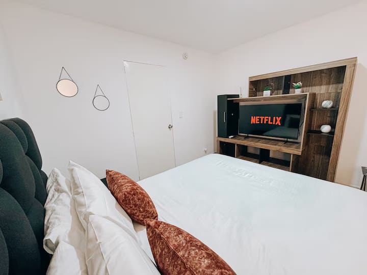 Acogedora habitación con cama doble, cabecera acolchada, cojines en tonos terracota y blanco. Frente a la cama, un mueble de madera con TV y acceso a Netflix. Decoración minimalista con espejos decorativos y ambiente cálido y moderno.