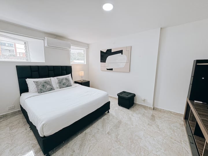 Habitación de estilo moderno con cama queen, cabecera acolchada negra y cojines decorativos. Espacio iluminado con aire acondicionado, lámpara de noche, puff, cuadro abstracto y mueble con TV. Piso claro tipo mármol y ventanas altas.