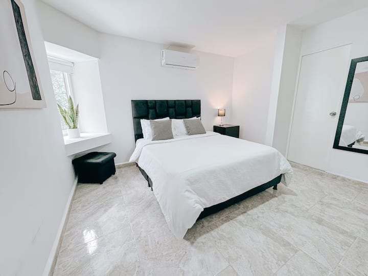 Habitación luminosa con cama queen y cabecera acolchada negra. Estilo moderno y minimalista con tonos neutros. Incluye aire acondicionado, espejo de cuerpo entero, lámpara de noche, puff, planta decorativa y piso claro tipo mármol.