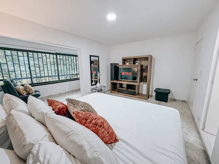 Amplia habitación con cama king, cojines decorativos en tonos tierra, sofá, espejo de cuerpo entero, centro de entretenimiento con TV y acceso a Netflix. Ventanal grande con reja y vista verde. Ambiente moderno, luminoso y acogedor.