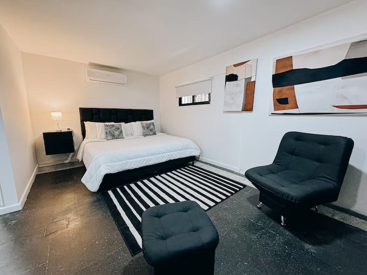 Habitación moderna con cama king, cabecero acolchado negro y aire acondicionado. Decorada con arte abstracto, alfombra a rayas en blanco y negro, dos sillas tipo lounge y mesa de noche con lámpara, perfecta para el descanso.