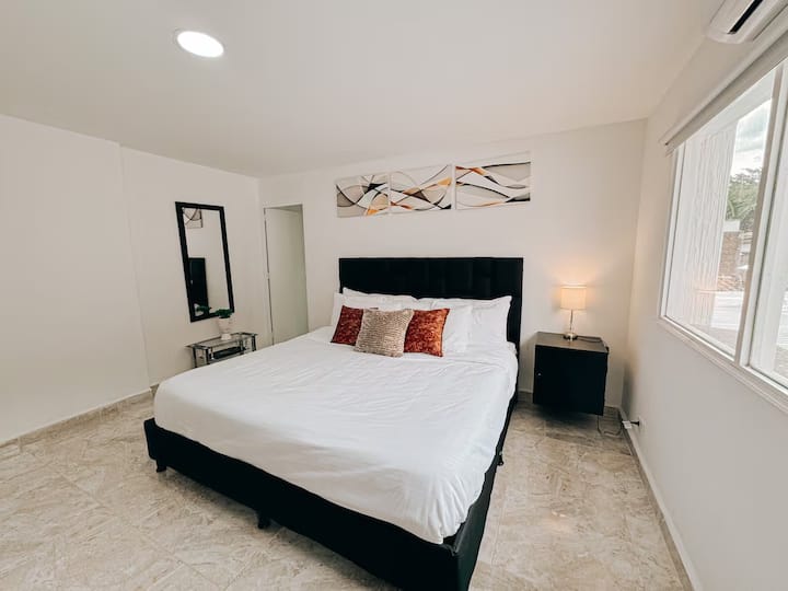 Moderna habitación con cama king, cabecera negra, cojines decorativos en tonos cálidos y ropa de cama blanca. Iluminación natural, lámpara de noche, espejo de cuerpo entero y arte abstracto sobre la cama completan un ambiente elegante y acogedor.