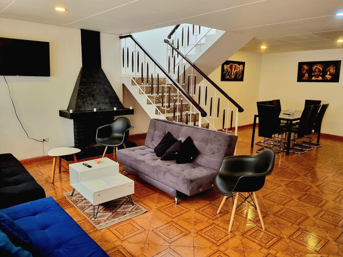 A spacious living area is depicted featuring grey sofas and chairs arranged around a white coffee table. A dining table with sleek black chairs is positioned nearby. A stone fireplace adds warmth to the setting, with a staircase leading to the upper floor in the background.