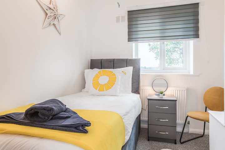 غرفة نوم فردية مريحة تتميز بسرير رمادي مع لمسات صفراء، ومنضدة سرير مطابقة، وسجادة فخمة. نافذة مع ستائر حمار وحشي تسمح بدخول الضوء. مسكنك الخاص الصغير لشخص واحد للاستمتاع بشكل مريح.