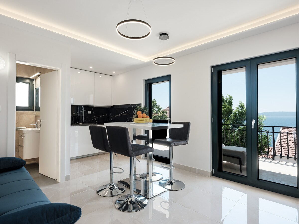 A modern open-concept kitchen and dining area is presented, featuring a sleek white cabinetry and a round dining table with high-backed chairs. Large glass doors provide access to a balcony with sea views, while a soft sofa is positioned nearby.