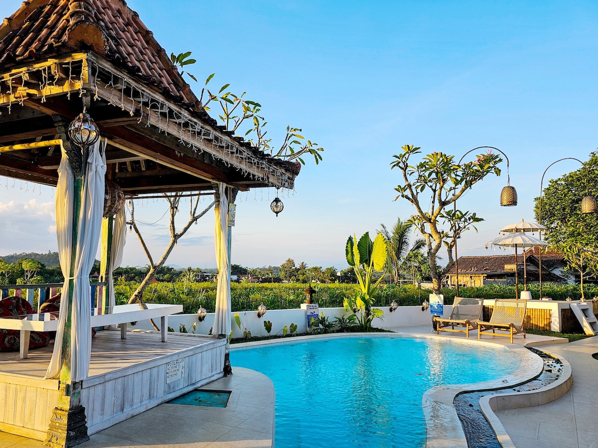 A gazebo with traditional architecture is surrounded by a small swimming pool, featuring clear blue water reflecting the sky. Tropical plants and trees enhance the natural setting, alongside loungers positioned for relaxation. The area is nestled among lush greenery, providing a serene ambiance.