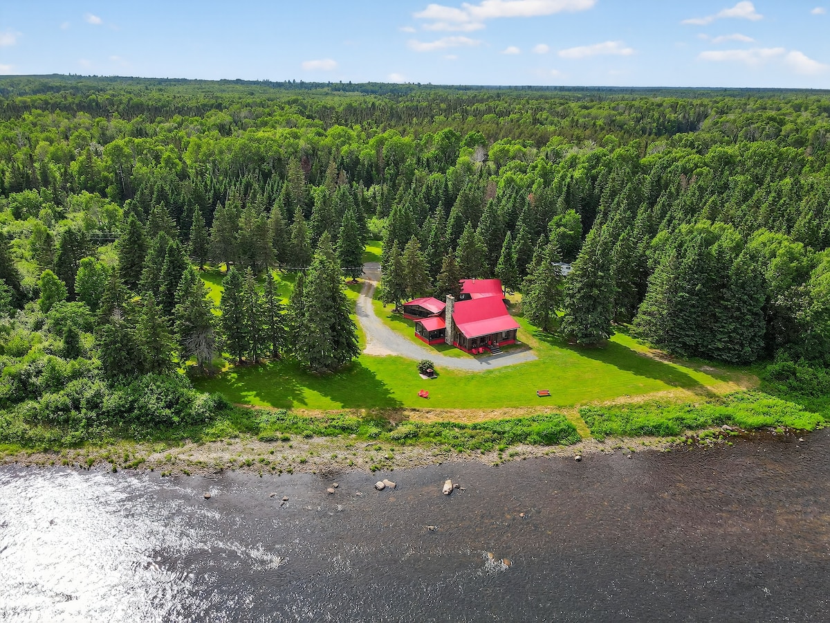 Aerial view of a secluded lodge nestled among lush greenery, with a vibrant red roof contrasting against the surrounding trees. A gentle river flows nearby, and a spacious lawn is visible, highlighting the tranquil natural setting.