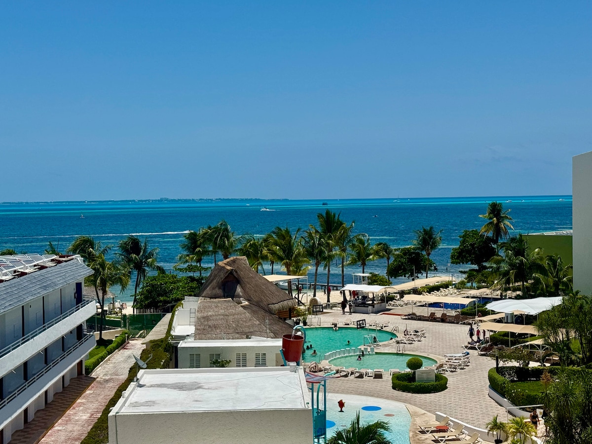 An expansive view reveals a spacious pool area surrounded by palm trees and lounge chairs, set against a backdrop of the ocean. The tranquil turquoise water meets a clear blue sky, with beach umbrellas providing shaded relaxation spots nearby.