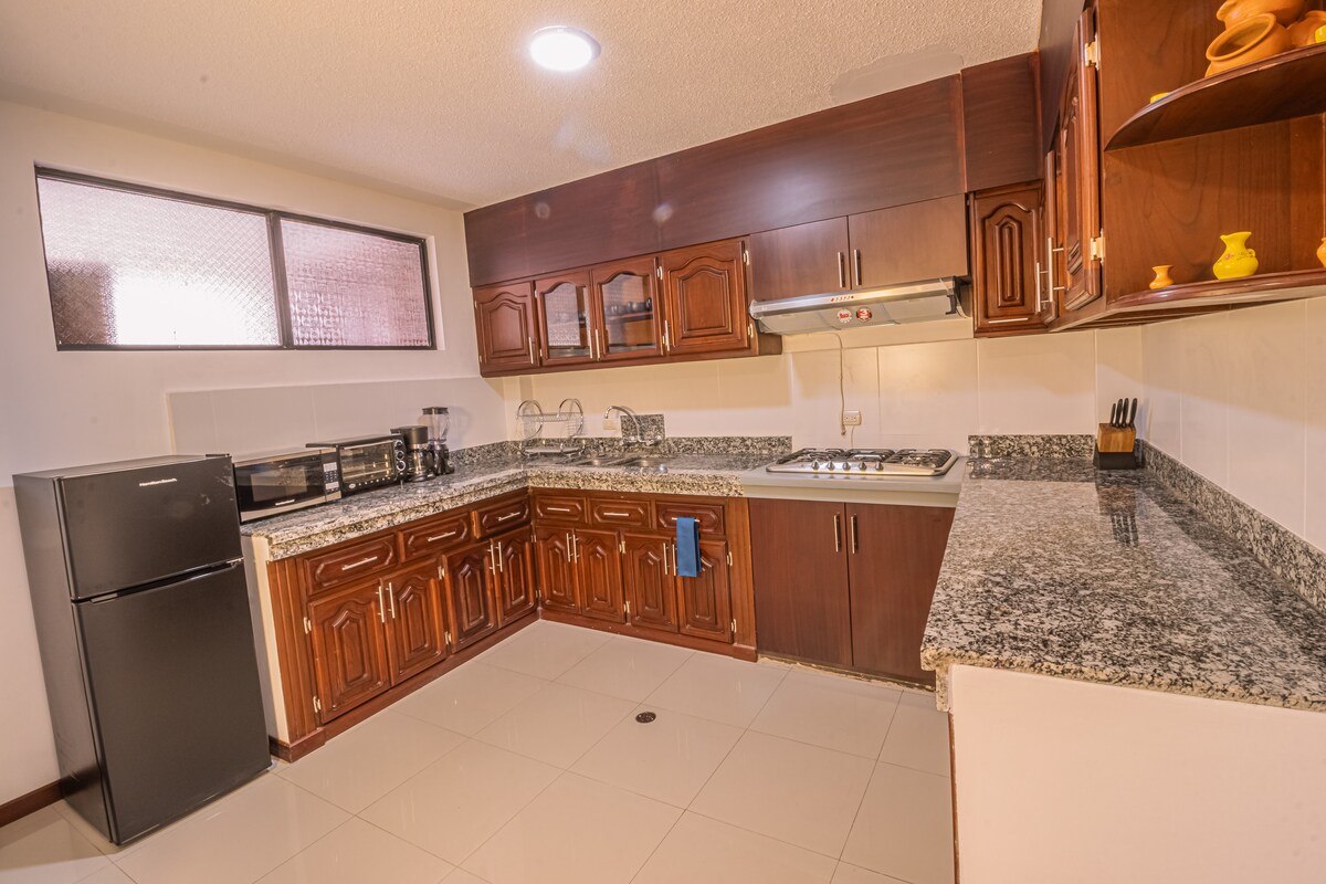 The kitchen features wooden cabinetry with a granite countertop and modern appliances including a refrigerator, microwave, and stove. Natural light enters through a window, enhancing the functional layout designed for cooking and dining.