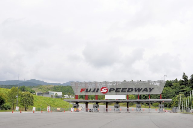 The entrance to Fuji Speedway is shown, featuring a modern architectural design with a large 'FUJI SPEEDWAY' sign prominently displayed. Surrounding greenery and hills create a natural backdrop, while a clear sky with soft clouds is visible above.