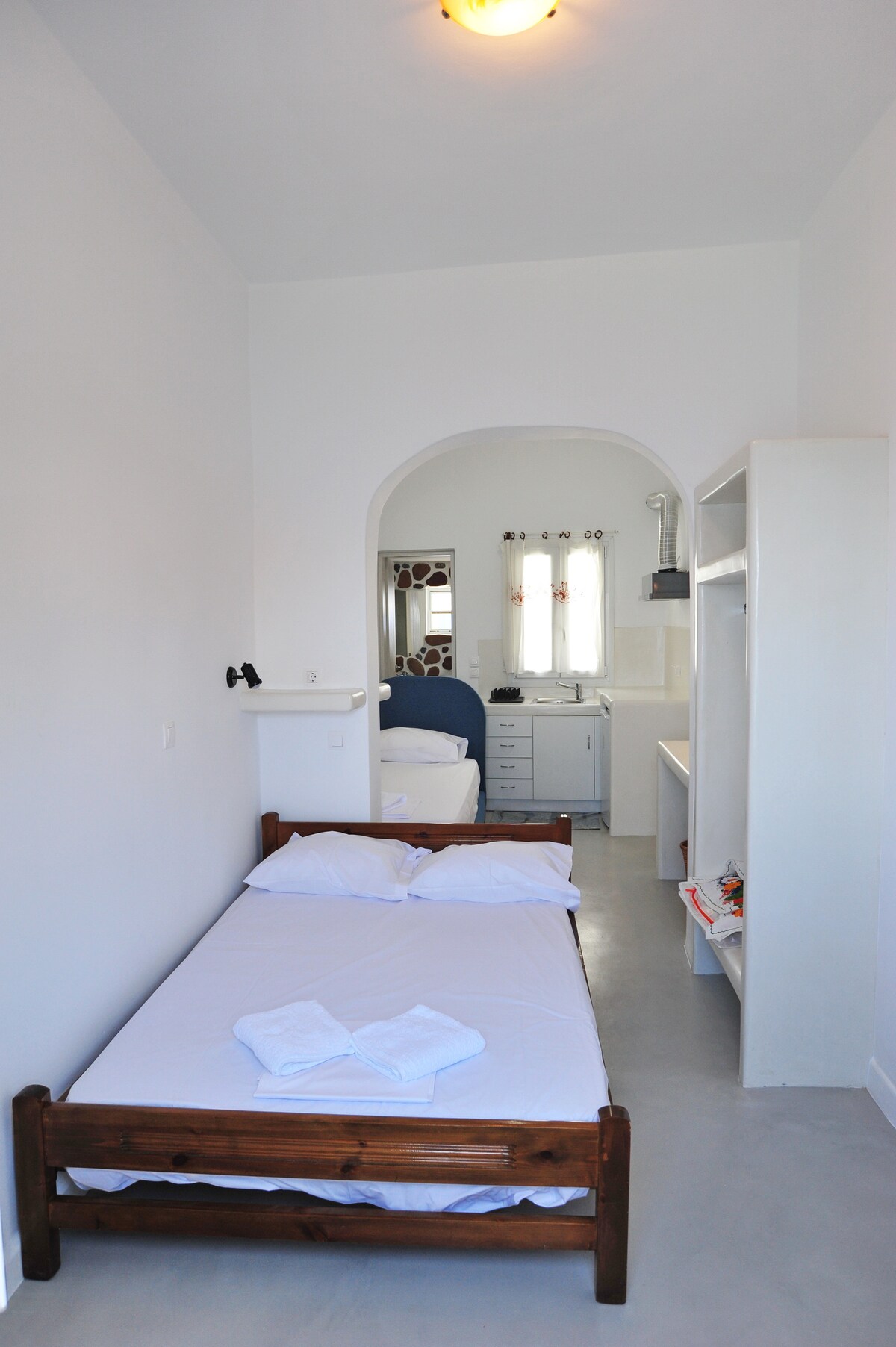 A bright studio space is presented, featuring a cozy sleeping area with a wooden bed and fresh linens. A doorway leads to a kitchen and a second sleeping area, visible in the background. Natural light enters through a window, illuminating the neutral-toned walls.