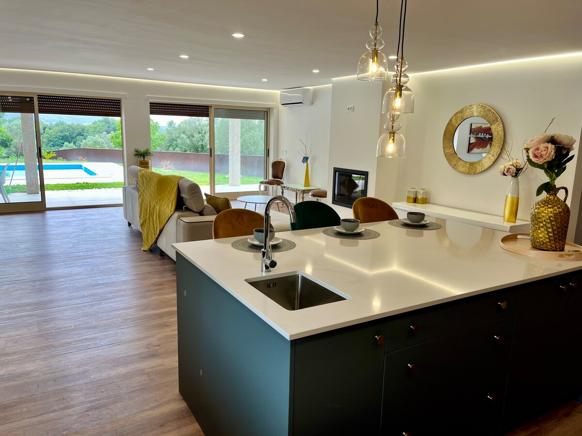 A modern kitchen features a central island with a sink and dining setup. Soft lighting is provided by pendant fixtures hanging above, while a view reveals large glass doors leading to a patio and swimming pool area beyond. Neutral tones and a cozy ambiance are evident.