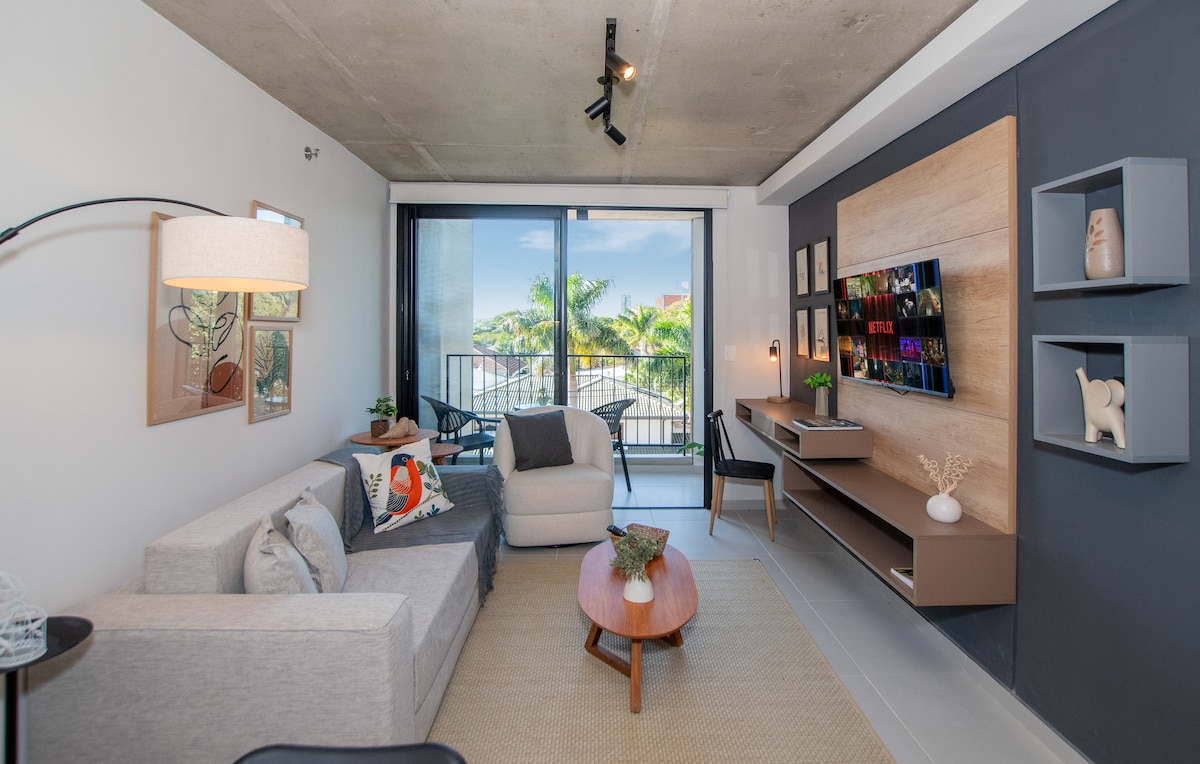 A modern living area features a light gray sofa and a wooden coffee table, complemented by decorative elements. Large glass doors lead to a private balcony with views of palm trees. A mounted smart TV and shelves add functionality and style to the space.
