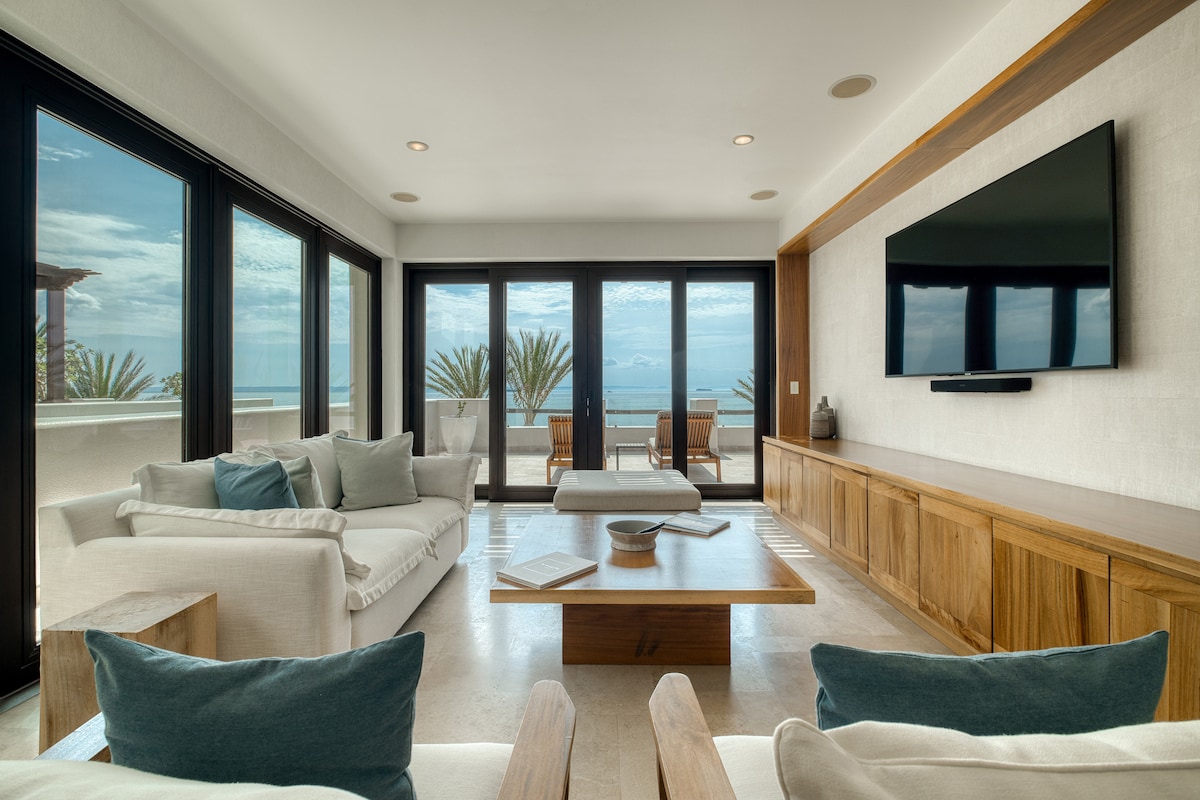 A bright living room features large glass doors that provide views of the ocean. A spacious white sofa is complemented by cushions in soft blues and greys. A wooden coffee table sits center, with decorative items placed atop. Natural light enhances the room's open and airy feel.