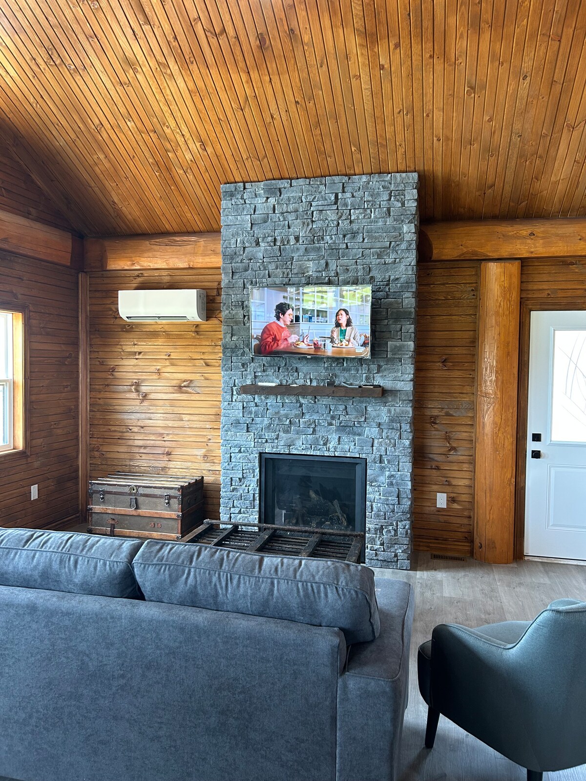 A cozy living area features a large gray sofa facing a stone fireplace, with a mounted television above. The warm wooden ceiling creates an inviting atmosphere. An air conditioning unit is visible on the wall, and a wooden chest adds to the room's functionality.