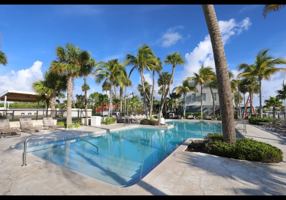 A clear swimming pool is surrounded by palm trees, with lounge chairs positioned along the deck. The pool features a gradual entry, and the sky is bright blue, dotted with fluffy white clouds, creating a relaxed and inviting outdoor setting.