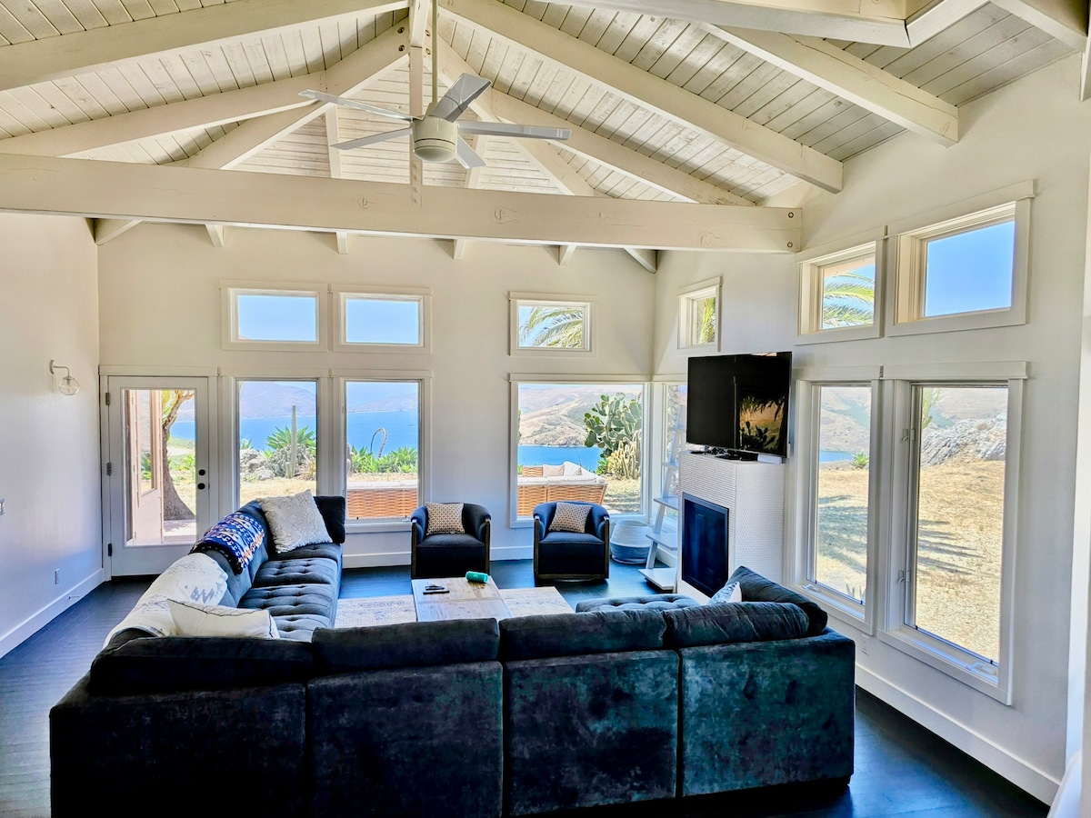 An open living area features a large sectional sofa, two armchairs, and a coffee table. Large windows provide unobstructed views of the surrounding landscape. A TV is mounted on the wall, and a modern ceiling fan enhances the airy atmosphere.