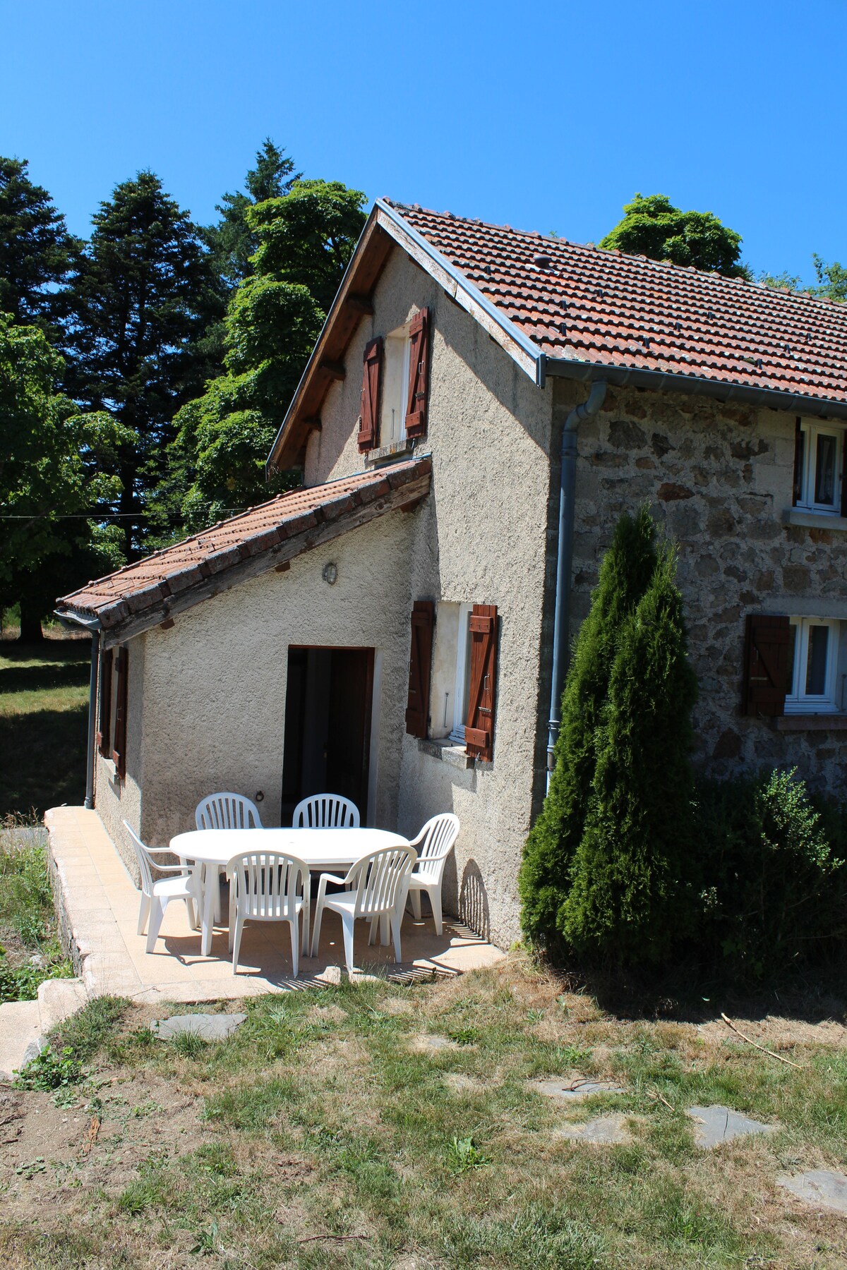 The exterior of the maisonette features a charming entrance with a textured wall and wooden shutters. A patio area is highlighted, furnished with a white table and matching chairs, set against a backdrop of lush greenery under clear blue skies.