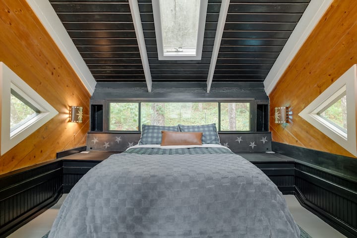 El dormitorio del loft cuenta con una cama de tamaño completo incorporada a medida con mesitas de noche, vista al jardín trasero desde los ventanales detrás de la cabecera y ventanas triangulares únicas a cada lado. Observación de estrellas de temporada desde el tragaluz de arriba. 