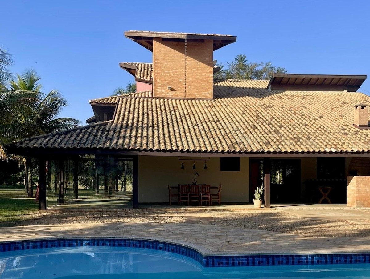 The exterior of the house features a spacious design with a tiled roof, surrounded by greenery. A swimming pool is visible in the foreground, with a dining area shaded under a roof. Large windows allow for natural light to enter the interior.
