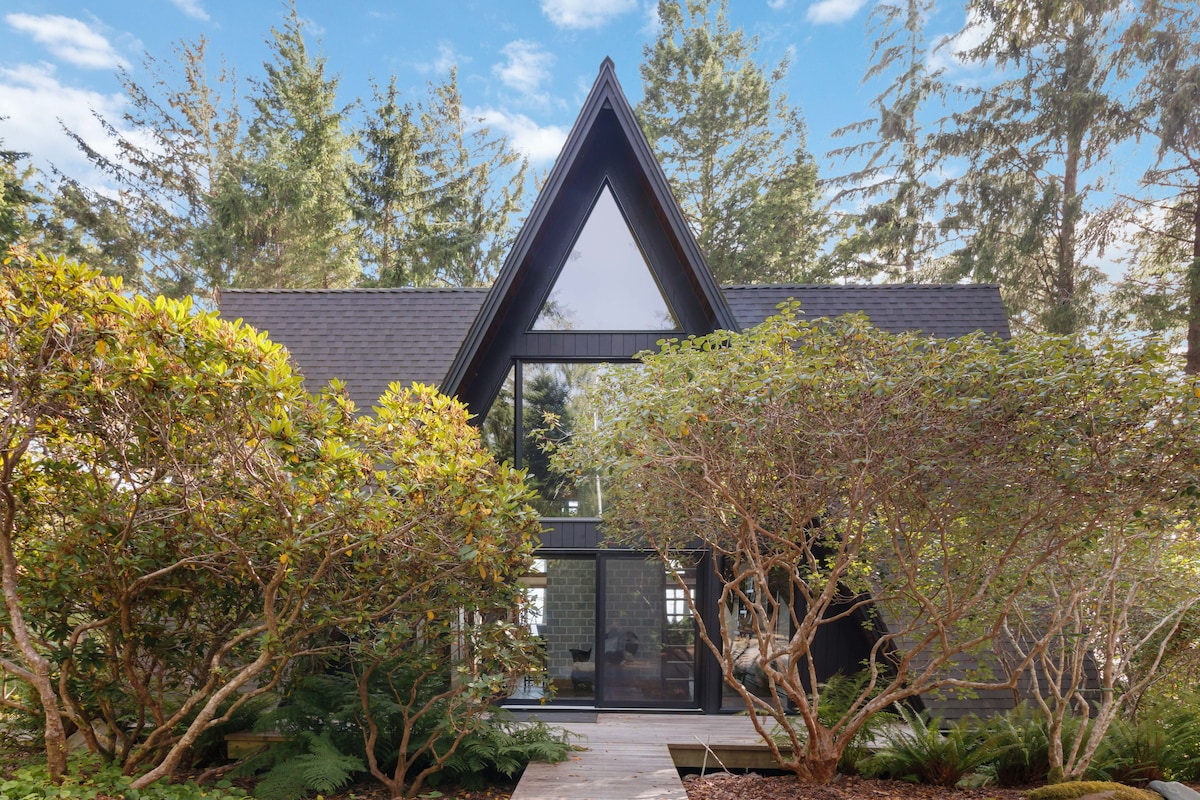 A modern A-frame cabin is framed by lush greenery, with floor-to-ceiling windows reflecting the surrounding forest. The structure showcases contrasting dark wood finishes against the natural landscape, creating a serene entrance. A pathway leads to the inviting front door, inviting guests to explore inside.