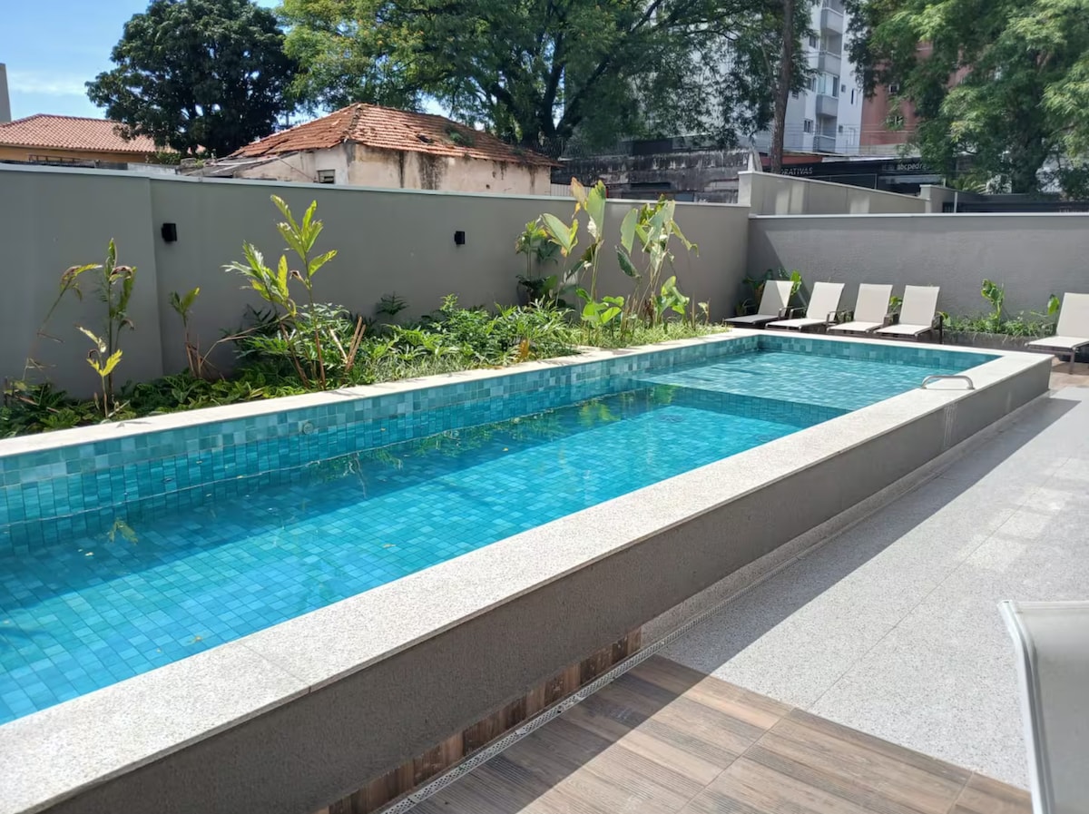 A modern swimming pool is shown, framed by lush greenery and contemporary loungers along the side. The pool features a clear blue tile surface reflecting the sky, while the surrounding area is designed with clean lines and a minimalist aesthetic.