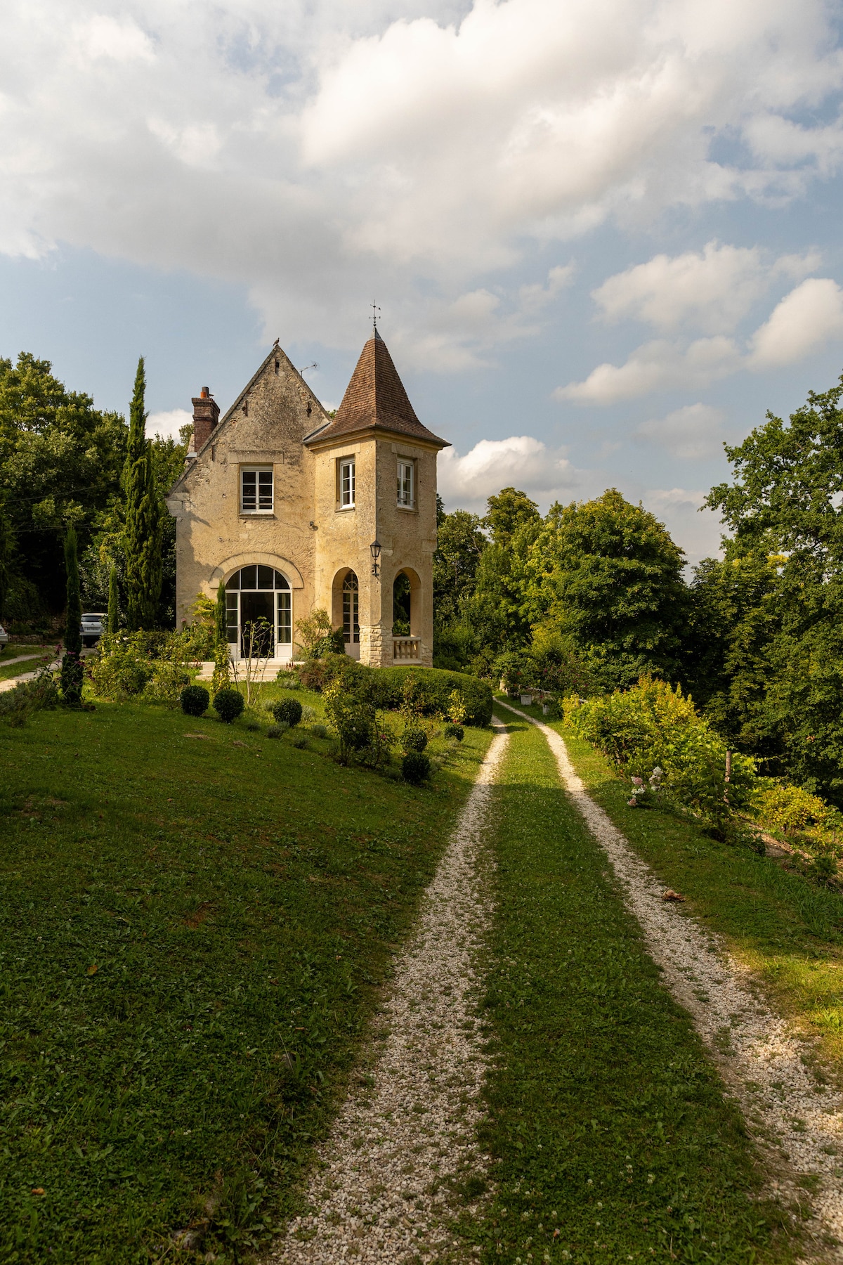 The quaint chateau is framed by lush greenery, showcasing a charming stone exterior and a distinctive tower. A gravel pathway leads to the entrance, flanked by manicured bushes, inviting exploration of the surrounding natural beauty.