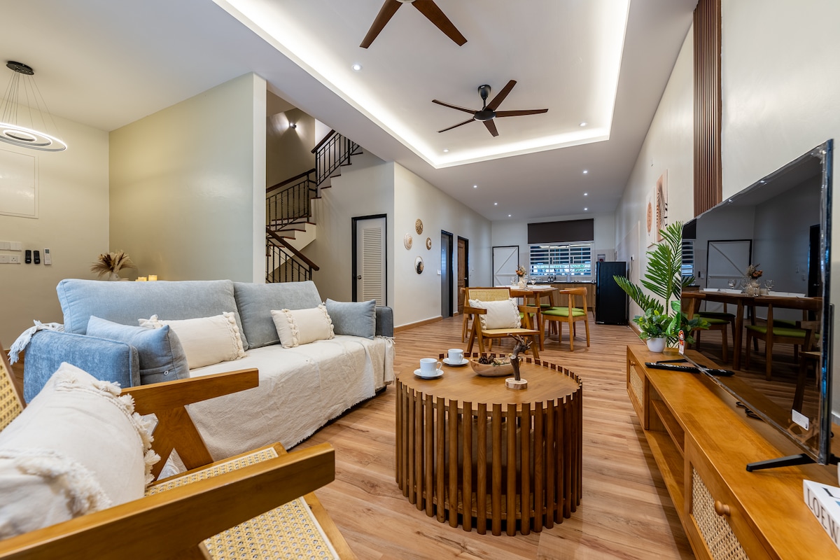 A spacious living area showcases a soft blue sofa paired with wooden chairs. Natural light fills the room, highlighting the circular wooden coffee table at the center. A TV unit and decorative plants add to the relaxation space, which flows smoothly into the dining area.