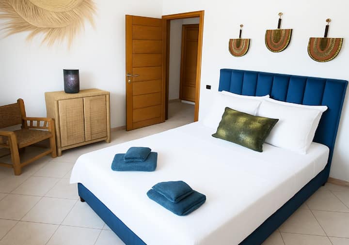 Svijetla spavaća soba koja nudi čist ambijent s bračnim krevetom, bijelom posteljinom i namotanih ručnika. Uređenje je inspirirano marokanskim majstorskim umijećem s namještajem od ratana i prirodnim zidnim predmetom. 