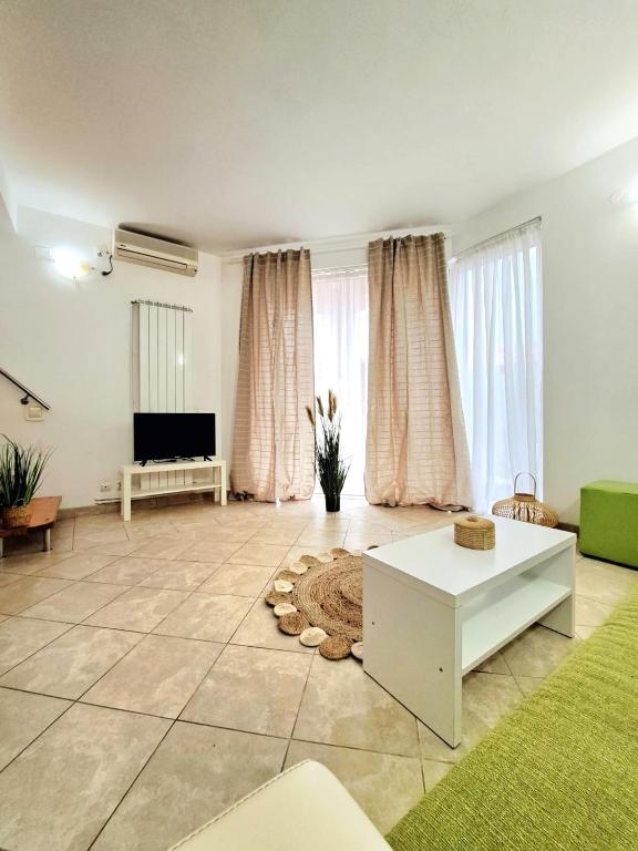 The living room is highlighted by soft natural light flowing through large windows, framed by light curtains. A small TV is positioned on a white stand, facing a simple coffee table. Textures and colors are introduced through a green sofa and a cozy rug beneath.