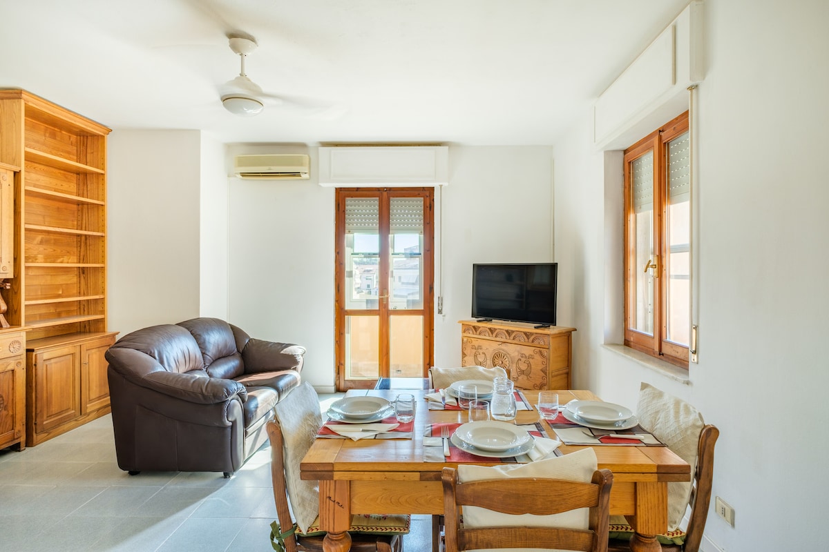 The living area features a spacious dining table set for a meal, accompanied by a comfortable brown sofa. Natural light fills the room through large windows with wooden frames, where a flat-screen TV is positioned against the wall, blending functionality with a relaxed atmosphere.