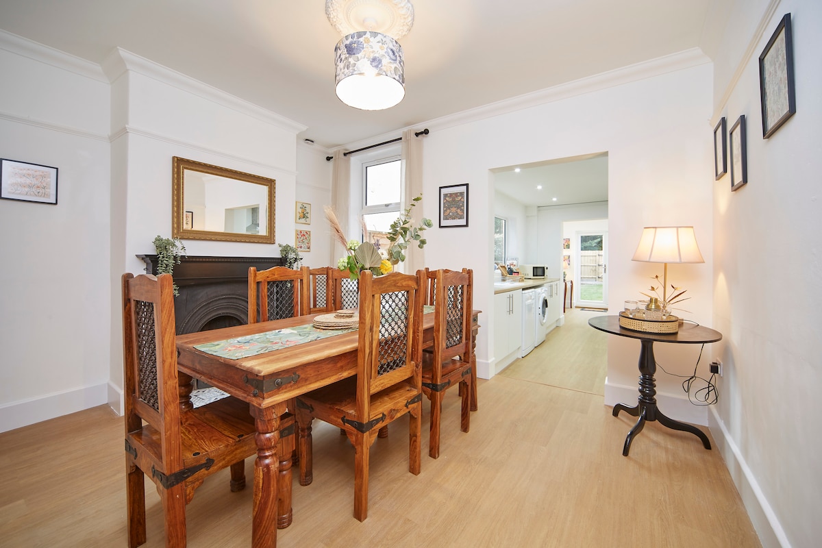 The open plan dining area features a wooden table that seats six, complemented by traditional-style chairs. Soft light is provided by a ceiling fixture, and a mirror enhances the sense of space. A doorway leads to the well-equipped kitchen in the background.