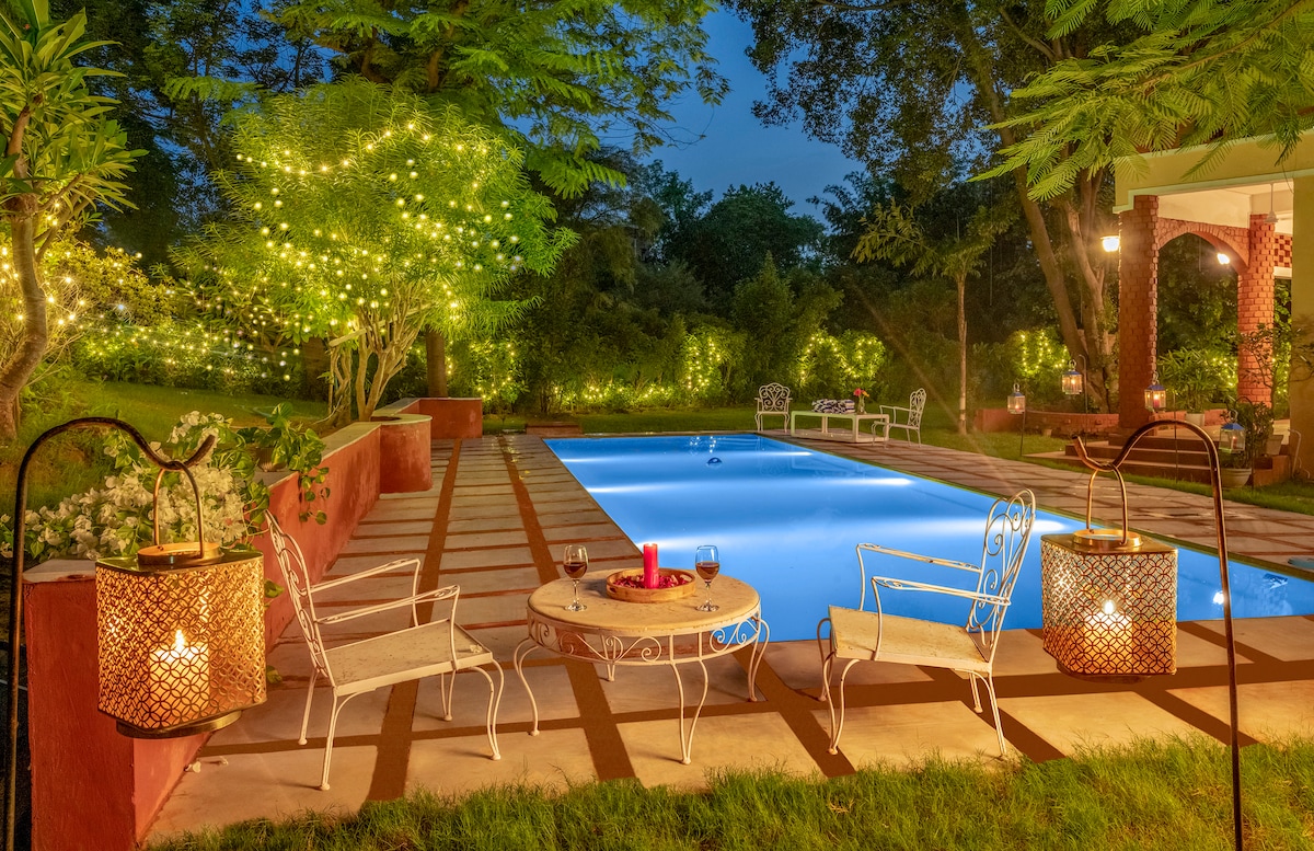 A serene outdoor area showcases a swimming pool surrounded by lush greenery. Elegant white chairs are positioned near the pool, with decorative lanterns providing soft lighting. Twinkling string lights are draped along the nearby trees, enhancing the tranquil ambiance.