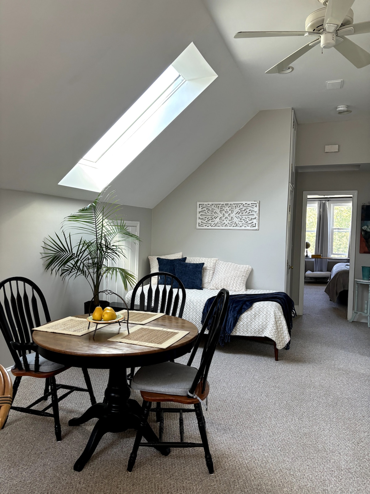 A comfortable living space showcases a round wooden table surrounded by four chairs. A potted plant adds a touch of greenery. Natural light filters through a skylight above, illuminating the area. A spacious bed is visible in the background, complemented by soft bedding.
